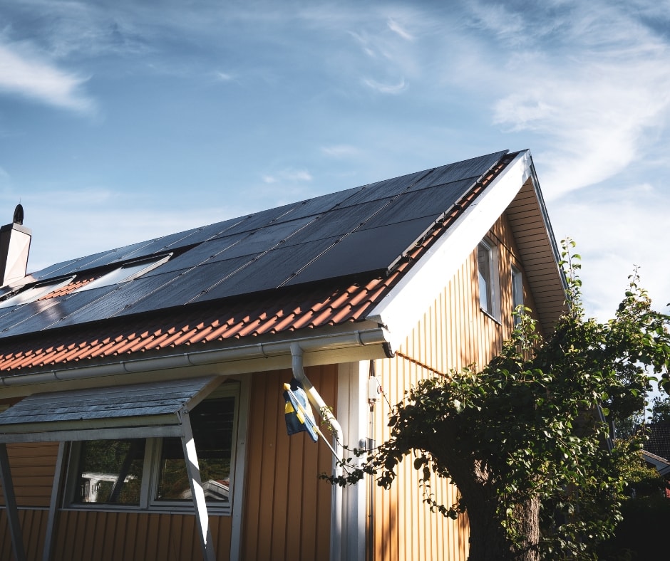 Svarta solceller som producerar grön el på gul villas tak, blå himmel med slöjmoln i bakgrunden och äppelträd i förgrunden.