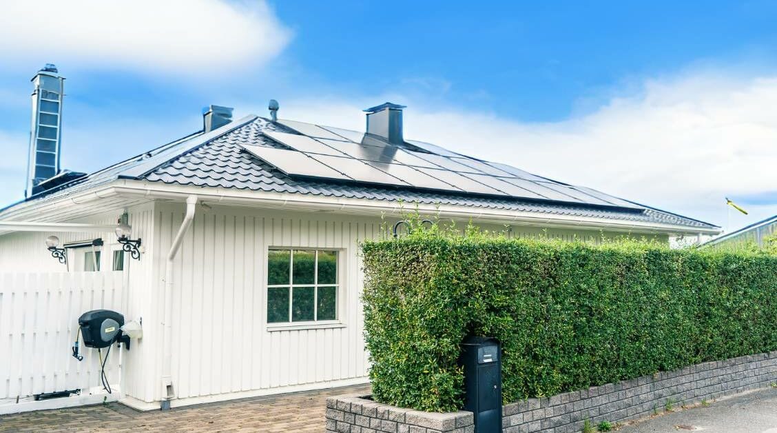 Vit villa med solceller på svart tak. Grön häck i förgrunden och blå himmel med slöjmoln i bakgrunden