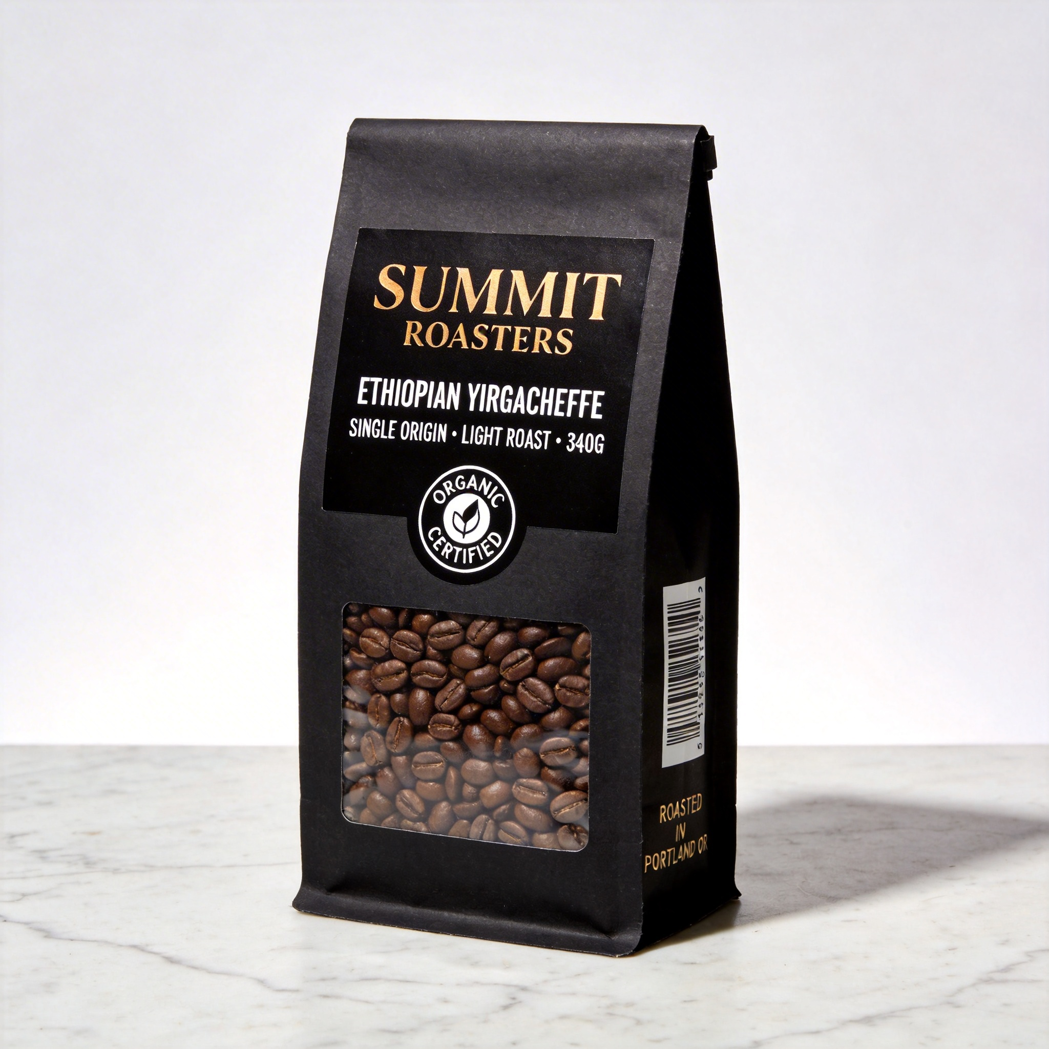 Seedream 4.5 - Product photography of a premium coffee bag standing upright on a marble countertop, the bag made of matte black kraft paper with a clear window showing whole beans inside, the front label reading SUMMIT ROASTERS in gold foil embossed serif font at the top, below that ETHIOPIAN YIRGACHEFFE in smaller white sans-serif, below that SINGLE ORIGIN * LIGHT ROAST * 340G in even smaller caps, a circular certification stamp reading ORGANIC CERTIFIED in a ring around a leaf icon, on the side of the bag a barcode and the text ROASTED IN PORTLAND OR