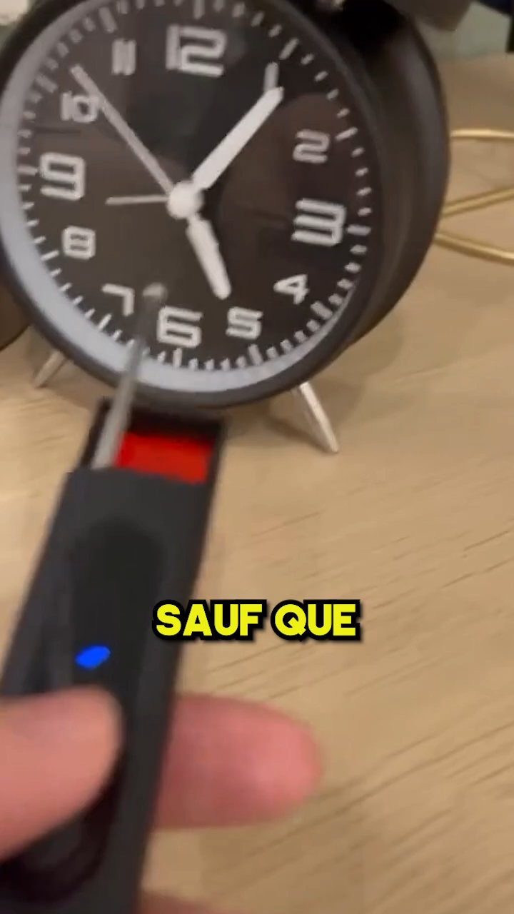 The image depicts a hand holding a black gadget with a button and a small antenna protruding from it, pointing towards an alarm clock in the background. The clock is displaying approximately 8:45. There is a large red arrow pointing to the antenna, suggesting it is an important focal point. The surrounding environment includes a lamp and other small items on a wooden surface, indicating this might be a bedside table.