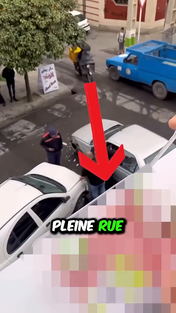 The image depicts an overhead view of a busy street where two white cars have collided. A man in a dark shirt and jeans stands next to one of the damaged cars, appearing to assess the situation. Another person in a uniform, possibly a uniformed officer, stands nearby. Several pedestrians and a motorcyclist are also visible in the background, observing the scene. The location seems to be an urban area characterized by the presence of trees and buildings.