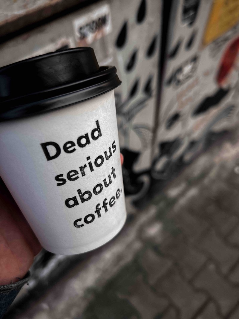 Картонена чаша с надпис Dead serious about coffee, държана на улицата пред изрисувана ролетка
