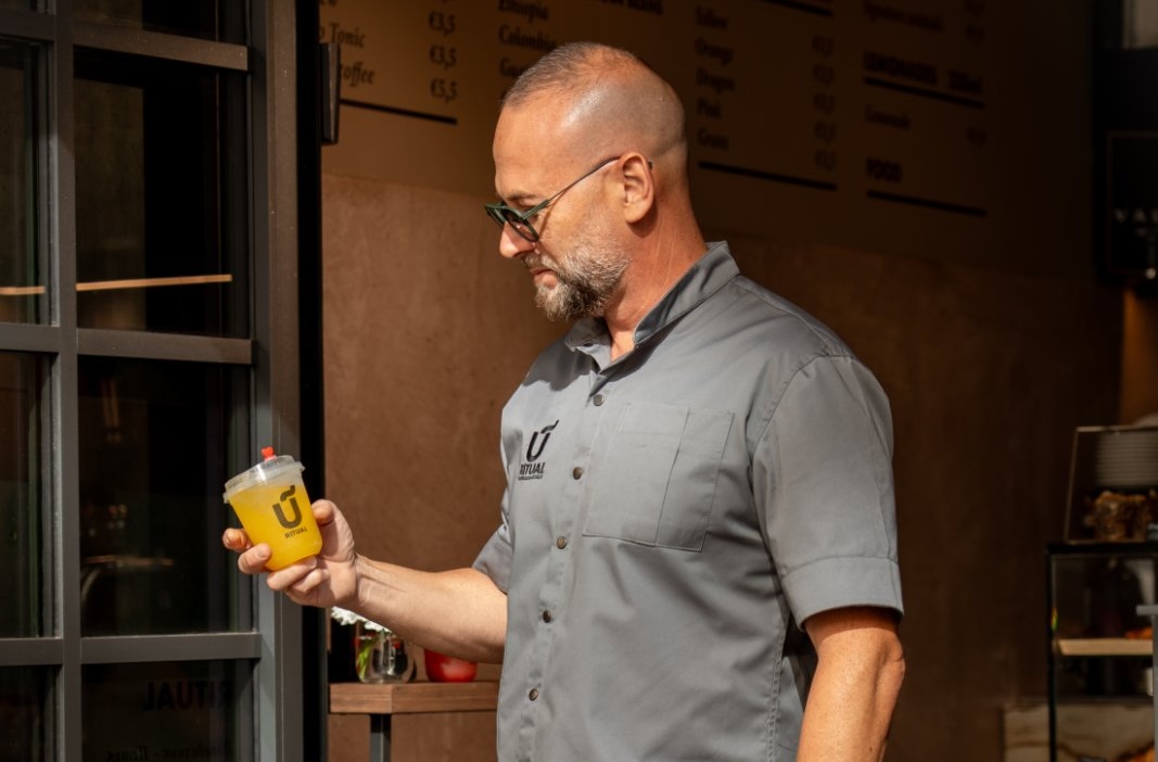 Dimitar Petrovski at the bar of Ritual Coffee and Cocktails, holding a yellow lemonade, the coffee menu on the wall behind him