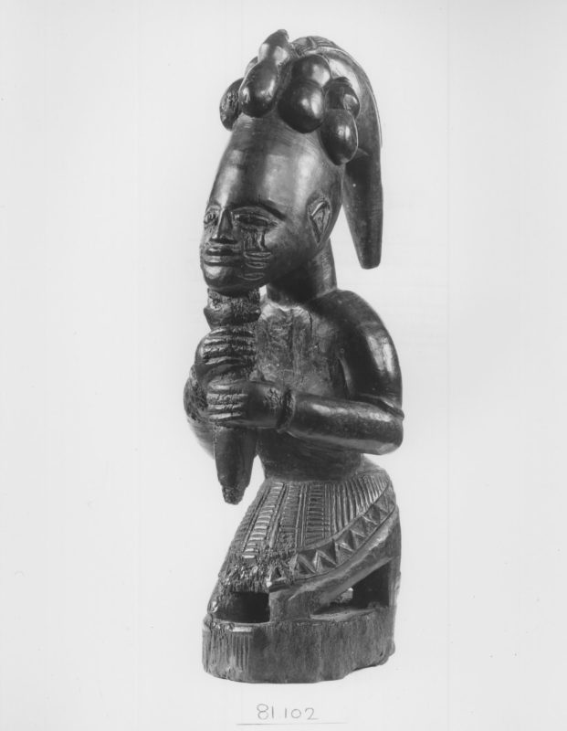 A photographic reproduction of a carved sculpture of a kneeling figure, Legba, the god of the crossroads, as well as the god of chaos; depicted here as a figure who serves as a connecting tissue between Africans on the continent and the Diaspora.