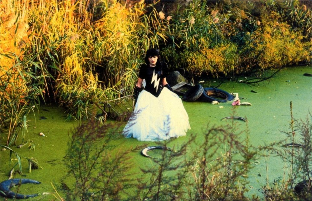 A light-skinned femme with long black long hair who’s wearing a white wedding dress with a black shirt with a thunderbolt on it. She is wading in a small green algae lake with abandoned tires and trash floating around her. There are tall bright yellow grasses encircling the lake.