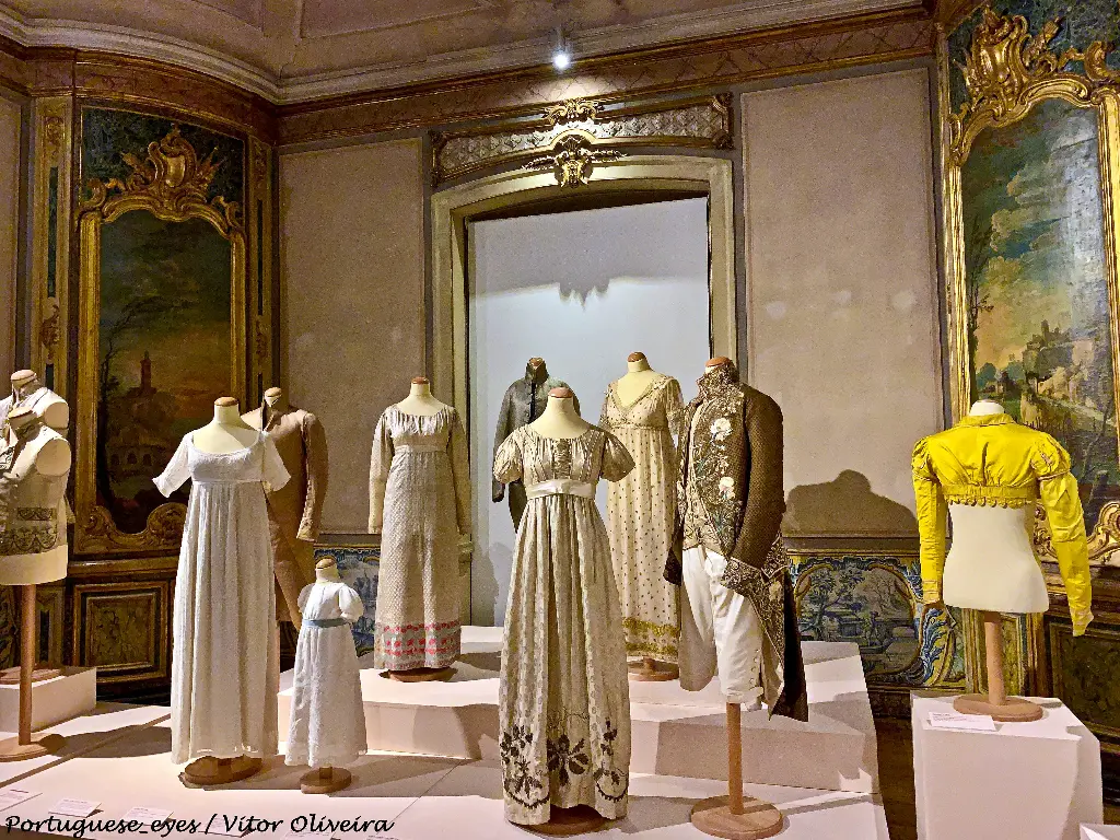 O Museu Nacional do Traje é o principal museu de âmbito nacional dedicado ao traje português e civil e traje estrangeiro - desde o século XVIII à atualidade.
O Museu está instalado numa vasta propriedade adquirida pelo Estado Português em 1975. Esta propriedade - uma antiga quinta de recreio setecentista - tem uma extensa área verde hoje aberta ao público e conhecida como - parque botânico do Monteiro-Mor, e a sua sede está instada num edifício setecentista - o Palácio Angeja-Palmela.
Coleções
O acervo da instituição reúne coleções de traje civil - feminino, masculino e de criança, nacional e internacional -, e respectivos acessórios, fragmentos de tecidos e peças de bragal, materiais e equipamentos que testemunham os processos de produção têxtil, traje e acessórios.
As primeiras ofertas de peças datam de 1974 nos registos do Museu Nacional do Traje, todas elas de particulares. A coleção pública que integrou o seu acervo veio do Museu Nacional dos Coches que, desde 1904, recolheu uma importante coleção de trajes da Casa Real.
Traje do século XVIII (estilos Barroco e Rocaille, 1700 a 1789)
Casaca, Séc. XVIII (c. 1770-1790)
No período Barroco, o traje feminino era geralmente composto por três peças principais, o corpete ajustado ao busto, a saia e a sobressaia. As mulheres vestiam-se sumptuosamente, sendo as rendas e os laços constantes no seu vestuário. Entre 1740 e 1770, em pleno período Rocaille, surge um vestido mais ligeiro e volante, com corpete, saia e o famoso "plis Watteau", composto por pregas de tecido que caíam soltas nas costas, sugerindo um falso manto. Em meados do século, o vestido “à francesa” utilizado nas grandes cerimónias, tinha amplos volumes laterais, um corpete cingido, e uma sobressaia aberta na frente, formando uma abertura triangular que deixava ver a saia.
A forma básica do traje masculino surgiu em França no final do século XVII, durante o reinado de Luís XIV, sendo composta por casaca, colete e calção. Este conjunto manteve-se no período Rocaille embora as casacas fossem menos amplas e se apresentassem ricamente bordadas. Os calções eram ajustados e terminavam abaixo dos joelhos. Os bordados formavam padrões florais e vegetalistas e eram realizados nas peças de tecido antes de se efetuarem os cortes quer das casacas, quer dos coletes.
Traje Império (1796 a 1820)
Casaca, colete e vestido Sec. XVIII-XIX
Com a Revolução Francesa, ocorrida em 1789, os ideais revolucionários de “Liberdade, Igualdade e Fraternidade” conjugados com um gosto pela Antiguidade Greco-Romana transformaram radicalmente o vestuário feminino. No traje feminino, as mulheres abandonaram os espartilhos e as anquinhas, assim como os pesados e ricos tecidos, os vestidos apresentavam linhas direitas, cintura subida e curtas mangas de balão, acompanhadas de luvas altas. As saias chegavam aos tornozelos e as caudas eram usadas apenas na corte.
Nas vésperas da Revolução, existia um grande entusiasmo pelas peças masculinas do traje inglês, quer pela qualidade da sua confeção quer pelo seu aspeto funcional. Mas a grande novidade desta época foi a introdução das calças no guarda-roupa masculino, provenientes do traje dos homens do povo e dos marinheiros, as calças começam a ser usadas como símbolo da revolução, pois os calções eram sinónimo do traje aristocrático.
Traje Romântico (1825 a 1865)
Vestido Romântico e Xaile, Sex XIX
No século XIX, época da industrialização, surgiram rápidos progressos tecnológicos em várias áreas da produção, aos quais a indústria da moda não foi alheia. Na década de 50 o traje feminino atingia a sua expressão máxima com a introdução da crinolina. Esta armação interna dava um grande volume simétrico às saias sem lhes adicionar peso. O espartilho voltou a moldar o busto feminino. Os tecidos preferidos eram as sedas e as musselinas de algodão com padrões de ramagens, flores, axadrezados e riscas. As cores eram simples e discretas, predominando os azuis e os verdes.
A moda masculina de 1850 manteve as tendências das décadas anteriores. Os casacos de cor preta ou de tons sóbrios usavam-se também com calças aos quadrados. Para a noite usava-se a casaca preta com calças e colete do mesmo tecido, camisa com peitilho engomado e laço.
Traje Belle Époque (1870 a 1914)
Saia e Casaco, Portugal, Sec. XX
As senhoras usavam vestidos de duas peças, compostos por corpo e saia, que eram confecionados com tecidos pesados e o espartilho manteve-se em voga. A saia usava-se comprida, acumulando drapeados, passamanarias, berloques, fitas, laços, pompons e franjas. Contudo, a característica silhueta deste período era dada por uma volumosa armação interior, chamada tournure, aplicada na parte detrás da saia.
Em 1890, as mulheres viram surgir os espartilhos ditos "saudáveis" que produziam um porte ondulado em forma de "S". Desta forma o busto da mulher ficava erguido e tão realçado que se denominava "peito de rola". As saias compridas em forma de campânula exibiam, geralmente, uma pequena cauda. Os vestidos de dia tinham um cós de pescoço alto e um peitilho de renda ou tule. À noite os vestidos apresentavam decotes amplos e os braços eram protegidos com luvas compridas.
No Traje masculino, as sobrecasacas e os fraques continuavam a ser usados em situações de maior cerimónia, com cartola. Para o quotidiano os conjuntos de três peças – casaco, colete e calças – eram usados com chapéu de coco.

Vestido Anos 20
Traje do século XX
(1910-2000)
A República implantada em Portugal a 5 de Outubro de 1910 coloca um fim à monarquia e transforma a dinâmica social da vida portuguesa. Muitos formalismos e constrangimentos sociais desapareceram e as artes, assim como a moda, foram palco de várias mudanças. Os desportos e as atividades ao ar livre promoviam um novo estilo de vida e as senhoras preferiam trajes de corte masculino, como o tailleur. De origem inglesa, o tailleur formado por saia e casaco adaptava-se bem aos passeios e era especialmente apreciado pelas jovens que cada vez mais integravam o mercado laboral.
(1910-1918)

Casaca, Colete, Calças e Cartola &amp; Vestido Sec. XX (c. 1920)
As mulheres portuguesas aderiram rapidamente a uma nova silhueta estreita e esguia proposta em Paris, por Paul Poiret. Inspirado no traje do período napoleónico este costureiro propôs o uso de vestidos em substituição dos fatos femininos de duas peças até aí utilizados. Os seus vestidos apresentavam linhas direitas e cintura alta, o que possibilitava à mulher libertar-se do espartilho. Em Paris, os Ballets Russes de Diaghilev inspiravam a moda, trazendo influências orientalizantes para o vestuário através de novas formas, cores vivas e tecidos luxuosos.
O traje masculino manteve as formas do final do século passado. Nas ocasiões solenes usava-se o fraque e a casaca, no quotidiano a sobrecasaca começava a ser substituída pelo casaco curto, com colete e calças.
(1920-1930)

Vestido Anos 30
O vestuário feminino deu nesta década o passo definitivo para a funcionalidade, acompanhando o estilo Art Déco, caracterizado por uma decorativa e elegante geometria. Os vestidos de linhas direitas com cintura descaída, mostravam as pernas a partir dos joelhos. Os vestidos para dançar eram curtos com decotes redondos ou quadrados, habitualmente com saia em évasée e, por vezes, cobertos com vidrilhos, lantejoulas e missangas. As meias e os sapatos passaram a ser peças de relevo, enchendo-se de cores. As cores eram vivas, embora também se escolhesse o preto.
No traje masculino na década de 20, generalizou-se nas cidades o uso de casacos cintados de tweed, espinhado ou em xadrez, de tons sóbrios, acompanhados de calças largas com pregas e dobra na extremidade inferior, em tecido liso ou de fantasia. O smoking transformou-se definitivamente no traje masculino preferido para eventos semi-informais como jantares públicos ou privados, bailes e festas. De cor preta com lapelas de cetim de seda, era acompanhado com laço do mesmo tom.
(1930-1946)
Na década de 30 o traje feminino regressou a linhas mais curvilíneas, o comprimento da saia desceu e a cintura retomou a sua posição natural. Os vestidos de noite eram compridos, com costas desnudadas. Os tecidos mais apreciados eram os crepes e os cetins. As atrizes de cinema, com o seu glamour, inspiravam as mulheres e tornavam-se modelos a seguir. Madeleine Vionnet criou o corte em viés, técnica que dava às peças uma elasticidade e fluidez muito apreciadas, moldando os corpos femininos de forma sensual.
A Segunda Guerra Mundial (1939-1945) impôs grandes restrições materiais. O racionamento impunha peças de traje mais ajustadas e curtas. As mulheres vestiam tailleurs com cintura bem marcada, saia de linha direita, ombros enchumaçados e bolsos amplos, que davam à sua silhueta uma postura masculina, de inspiração quase militar.
(1947 a 1950)
Com o fim da Segunda Guerra Mundial, Christian Dior correspondeu aos anseios das mulheres criando uma silhueta feminina e luxuosa. O estilo proposto pelo costureiro francês foi denominado New-Look, pois surgiu como reação à funcional moda dos anos 40. Apresentado em Paris em 1947, pretendeu restituir à mulher o seu aspeto sensual. Esta nova imagem caracterizava-se pelo uso de saias direitas ou amplas, muito rodadas e armadas, cintura estreita e delicada, ombros pequenos e redondos, e corpo espartilhado com o peito bem definido.
Ao mesmo tempo que vigorava a aparência New Look, surgia uma geração inquieta, da classe média, que rejeitava o materialismo vigente. Nos Estados Unidos da América as t-shirts transformavam-se em peças exteriores, principalmente depois de Marlon Brando ter aparecido com uma t-shirt branca no filme "Há lodo no cais", em 1954. As calças de ganga, conhecidas como jeans, começaram a ser adotadas pelos jovens como expressão da sua rebelião.
(1960 a 1970)
Os Anos 60 representaram uma nova alteração no traje. A juventude tornou-se num modelo a seguir, veiculando uma atitude de inconformismo e de contestação à política e mentalidade vigentes. Os movimentos de moda partiram da rua e passaram a influenciar a Alta-Costura. Os fabricantes de roupa viram nos jovens potenciais clientes e começaram a criar peças especialmente para eles. Na base da moda feminina desta década destacou-se a minissaia, apresentada por André Courrèges, em 1965, e popularizada por Mary Quant.
No final desta década surgiu em São Francisco, na Califórnia, o movimento hippie que foi antes de mais um movimento de mentalidades e se difundiu através da música pop. O seu vestuário inspirava-se nos trajes étnicos internacionais: os rapazes vestiam túnicas de algodão cru e jeans, apresentavam cabelos compridos e barbas; as raparigas vestiam longas túnicas, usavam os cabelos soltos com flores e o rosto sem maquilhagem. 
(1980 a 2000)
As marcas de pronto-a-vestir passaram a ganhar importância com o surgimento dos estilistas que paralelamente à Alta-Costura, contribuíram para equilibrar a massificação advinda do desenvolvimento da indústria da moda. A relativa estabilidade e prosperidade dos anos 80 favoreceram, uma vez mais, o revivalismo de valores e gostos tradicionais. Todavia, os anos 90 configuraram-se a nível internacional, dando jus à aldeia global. A televisão, as revistas de moda, o turismo e a internet desfizeram as fronteiras, propiciando a divulgação das criações já não só francesas mas italianas, inglesas, japonesas e americanas.
As marcas de pronto-a-vestir desenvolveram-se a um ritmo crescente para responderem aos que desejavam veicular uma aparência contestatária. Por outro lado, surgem os designers de moda e o que veio a chamar-se "moda de autor", o que atribui para criar uma alternativa de qualidade e de estilo à massificação que o desenvolvimento da indústria têxtil e de confeção estavam operar.
História
Pedro de Sousa Holstein, Conde de Palmela
O Palácio Angeja-Palmela foi mandado erguer no século XVIII por D. Pedro José de Noronha, 3.º marquês de Angeja, próximo ao local onde existira o paço de D. Afonso Sanches, filho natural de Dinis I de Portugal (1279-1325).
Com traça de autoria desconhecida, o palácio apresenta influência da arquitetura pombalina, desenvolvendo-se em duas fachadas, uma das quais findando com a capela. Do primitivo paço subsiste apenas uma ombreira quinhentista em uma residência contígua ao palácio, e algumas estruturas arquitetónicas do século XVII.
A entrada principal do palácio desenvolve-se como uma galilé e a articulação entre os andares é feita por uma escada de quatro lanços retos. Nas suas salas destacam-se os tetos de masseira, os estuques, as pinturas ornamentais e diversos painéis de azulejos setecentistas.
Em 1840, o imóvel foi adquirido por D. Pedro de Sousa Holstein, marquês de Palmela e mais tarde 1.º duque de Palmela, que empreendeu obras de beneficiação do palácio, entre as quais a reconstrução do pavilhão neo-gótico, hoje ocupado pelo restaurante do museu.

Brasões dos Marqueses de Faial e 2º Duque de Palmela, Palácio Angeja-Palmela
A partir da Segunda Guerra Mundial (1939-1945), o palácio passou a funcionar como colégio religioso de belgas refugiadas, até que, em 1975, o Estado Português adquiriu a Quinta do Monteiro-Mor, que, para além do Palácio Angeja-Palmela, compreende o Palácio do Monteiro-Mor, uma residência do século XVIII, o Jardim Botânico e uma zona verde com onze hectares.
O Museu Nacional do Traje resultou de um projeto nascido em 1969, apresentado em 1973 e consolidado com a exposição O Traje Civil em Portugal apresentada no Museu Nacional do Arte Antiga em 1974. A responsável de todo este processo foi Natália Correia Guedes, que veio a ser a primeira diretora do Museu.
A 23 de dezembro de 1976 foi passado um Decreto/Lei instituiu o Museu Nacional do Traje e o Parque Botânico do Monteiro-Mor.
A 26 de julho de 1977, o museu foi inaugurado pelo então Secretário de Estado da Cultura, David Mourão-Ferreira, a diretora do museu Natália Correia Guedes, e a presença do então 1.º Ministro, Mário Soares, com a abertura de cinco exposições: História do traje civil e urbano (da Antiguidade a 1925), complementada com a exposição Trajo Popular (uma parceria com o Museu Nacional de Etnologia) e a que se somava ainda Traje de ópera, coleção de Tomás Alcaide. Exploravam-se também as Técnicas de fiação, tecelagem e estampagem e expunham-se Brinquedos dos séculos XVIII ao XX.
A biblioteca particular da museóloga Maria José de Mendonça encontra-se aqui depositada.

<a href="https://pt.wikipedia.org/wiki/Museu_Nacional_do_Traje" rel="noreferrer nofollow">pt.wikipedia.org/wiki/Museu_Nacional_do_Traje</a>