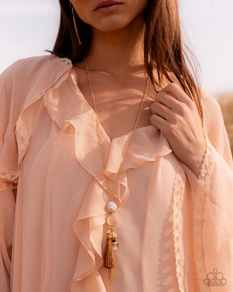 woman wearing light orange ruffled dress and a long pendant necklace featuring a white stone, gold t…