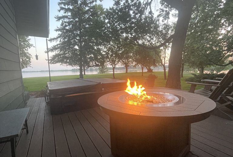 Fire pit table lit on an outdoor wooden deck overlooking trees and the lake at dusk