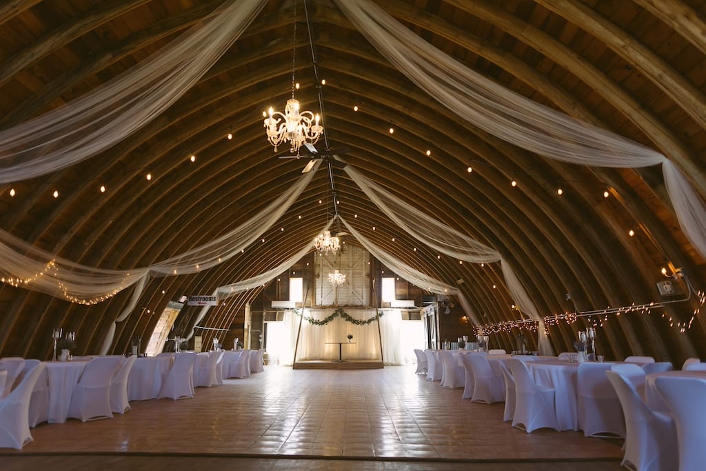 Wooden barn hall decorated with white fabric drapes, chandeliers, and reception tables