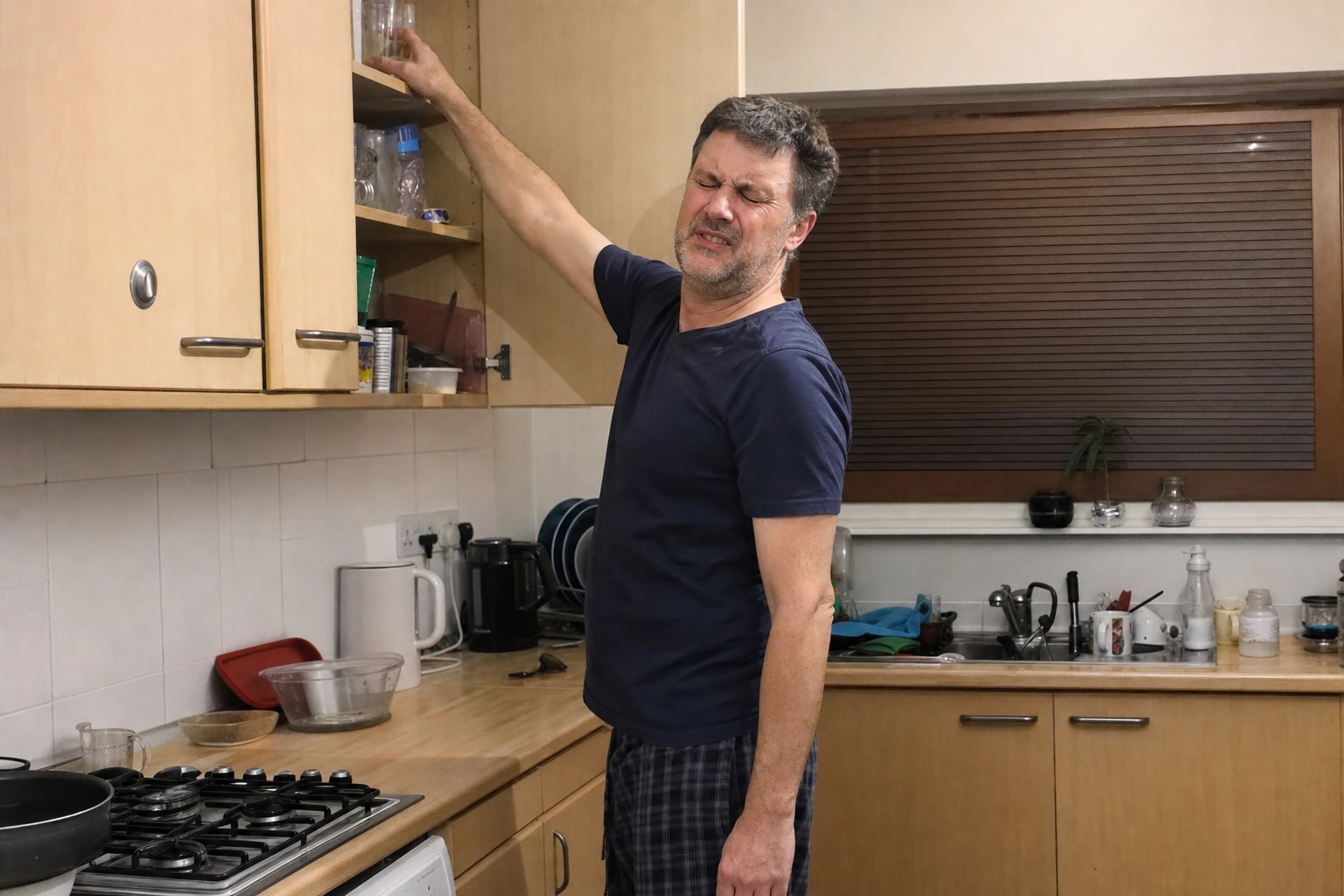 Man in his fifties wincing and gripping his shoulder as he reaches up to a kitchen cupboard