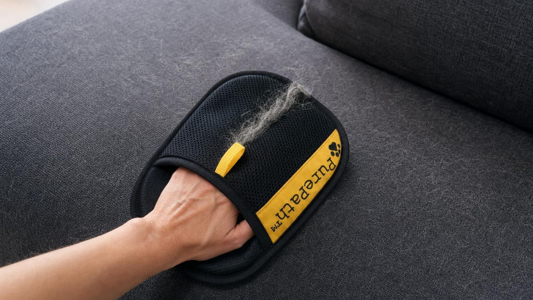 A hand wearing the PurePath glove pulling cat hair off a fabric sofa arm