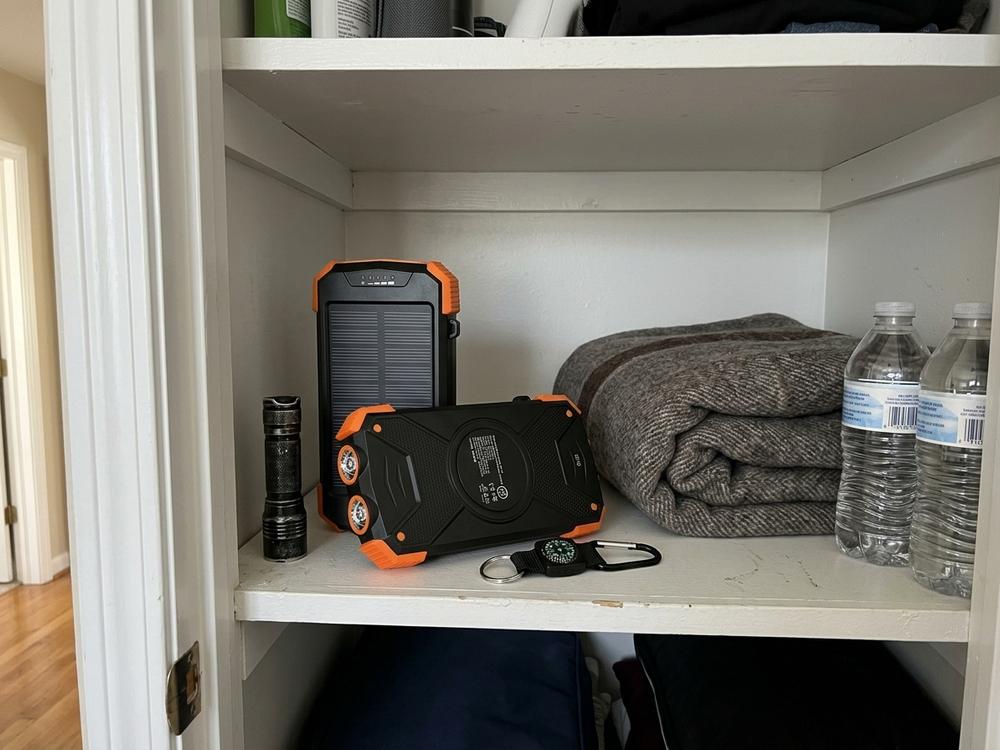 The power bank on a hall closet shelf beside a flashlight, a folded blanket and two bottles of water.
