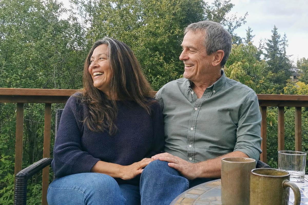 A long-married couple sitting close together on a back deck, his hand resting on her knee