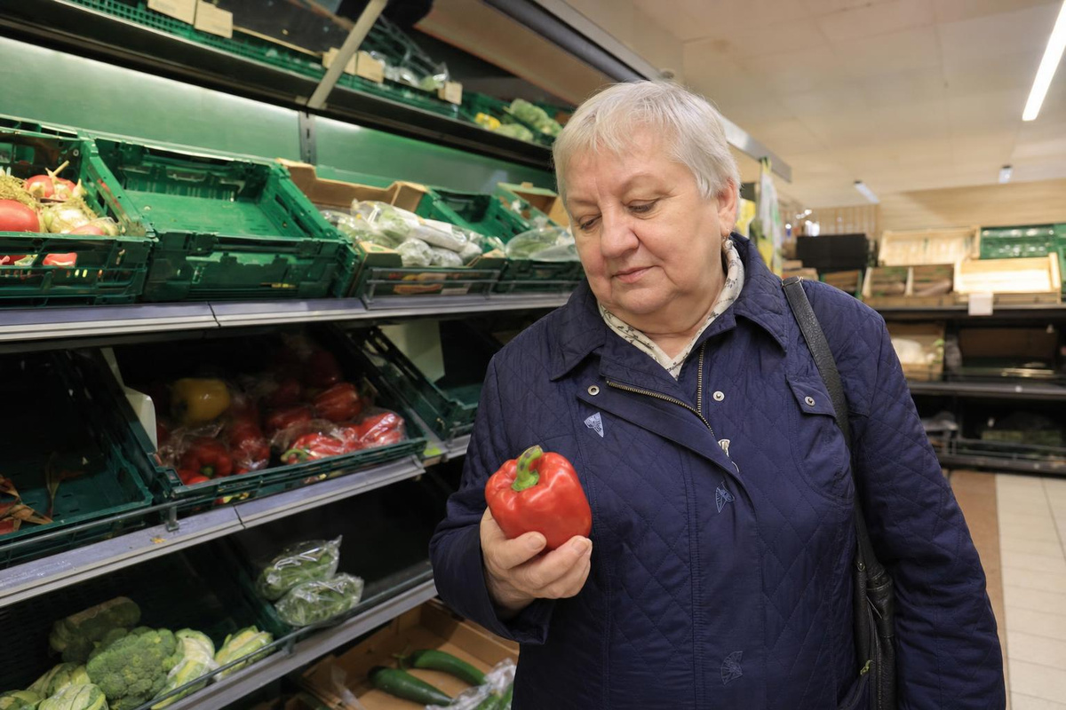 A woman in her late sixties in a supermarket produce section with sparse, picked-over bins, looking at a single item in her hand.