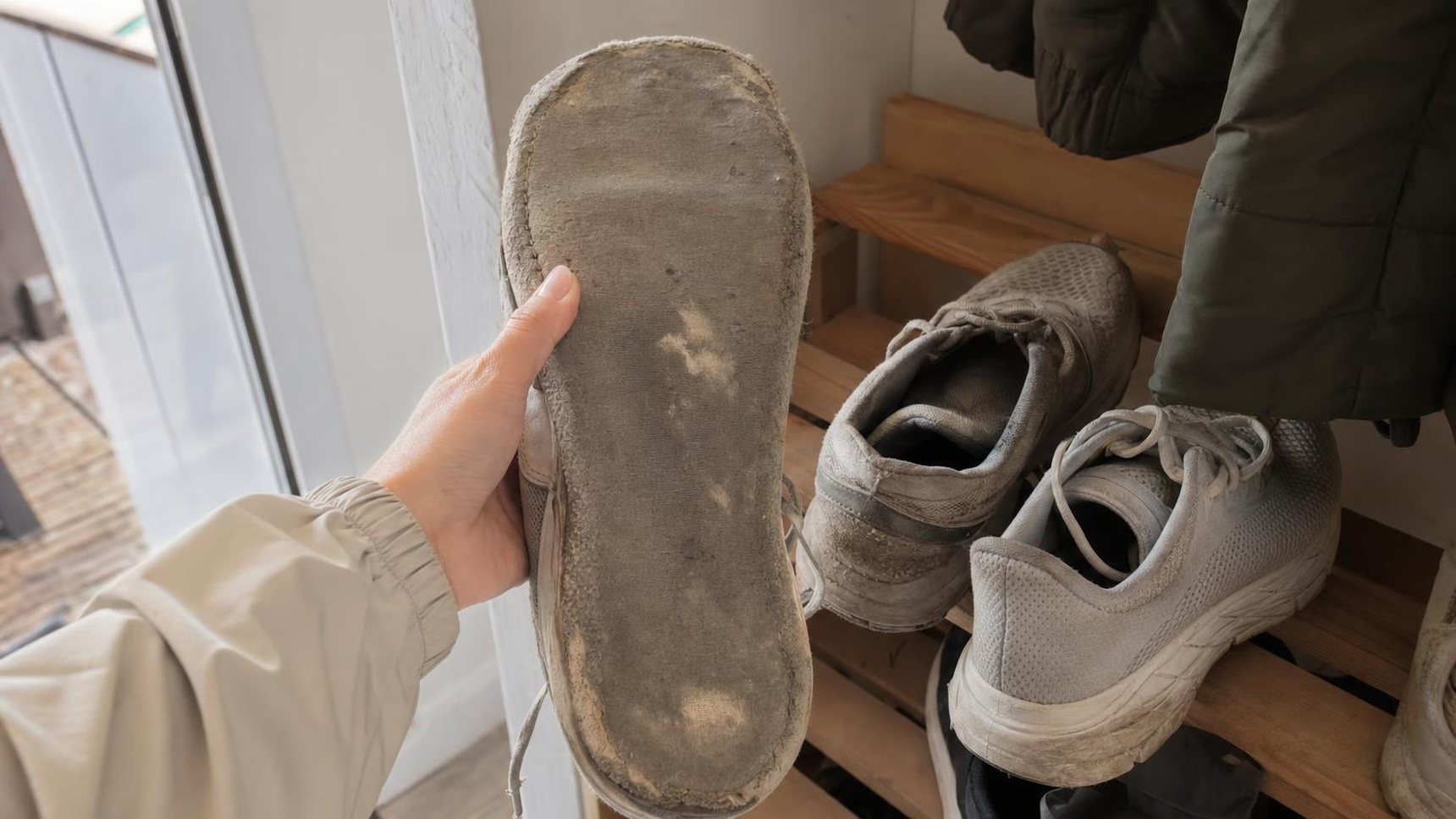 Una mano gira una vecchia scarpa da corsa per mostrare la suola consumata, accanto alla scarpiera di casa