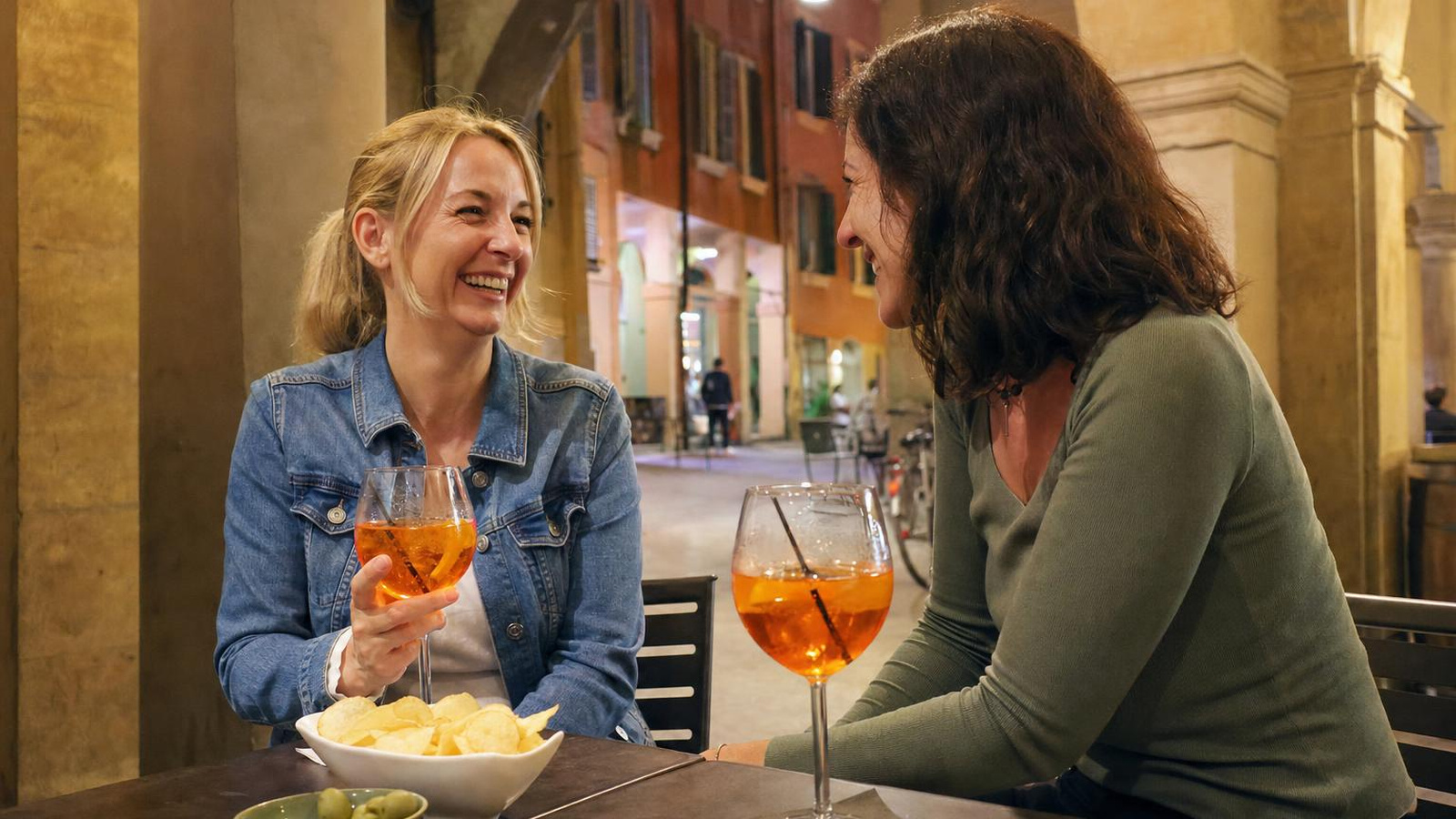 Due amiche ridono all'aperitivo, sedute a un tavolino all'aperto