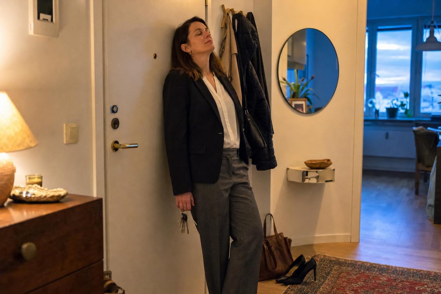 Donna appena rientrata a casa la sera, appoggiata alla porta con gli occhi chiusi, la borsa e le scarpe a terra