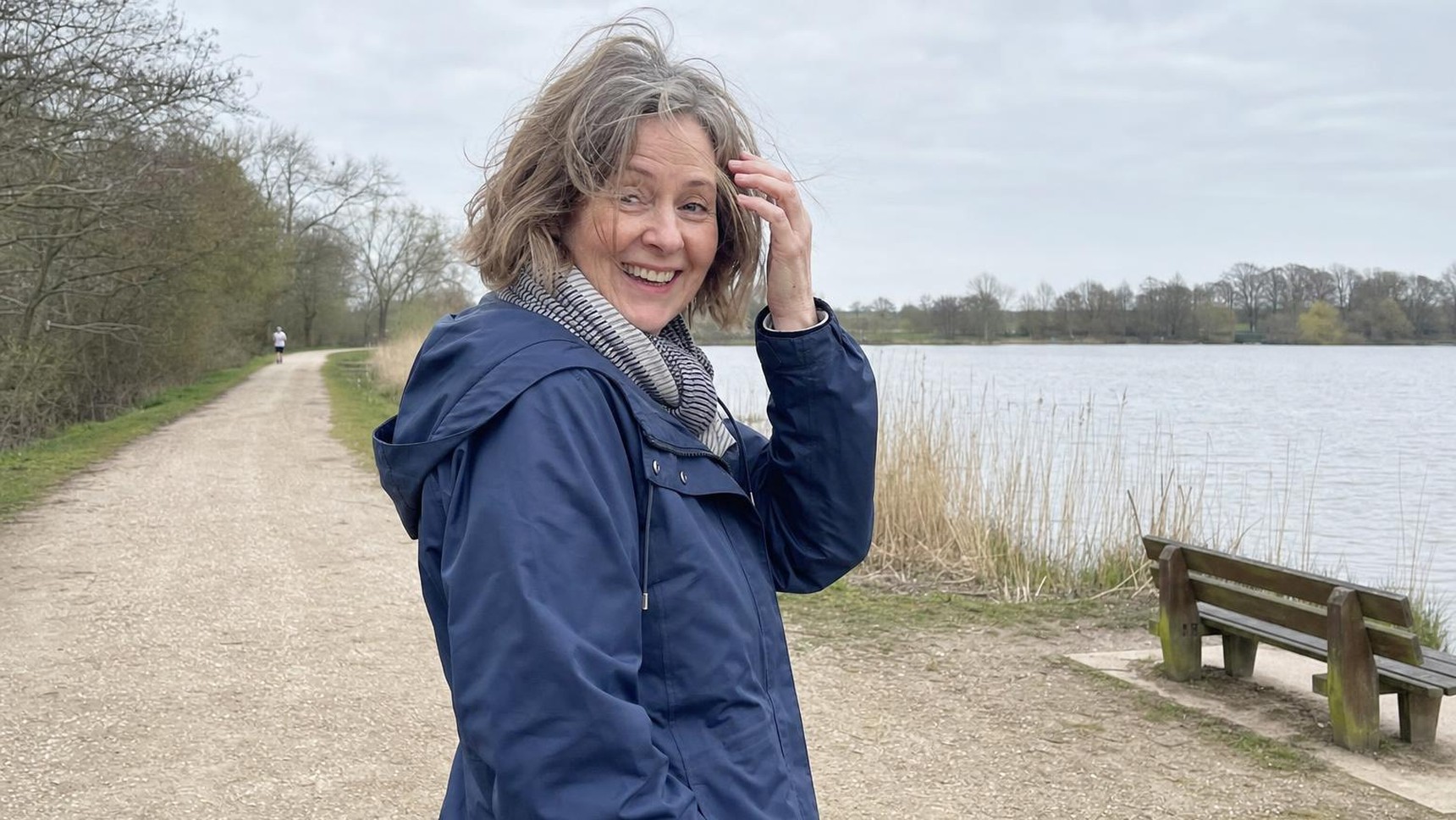 A woman in her fifties smiling on a lakeside walk on an overcast day
