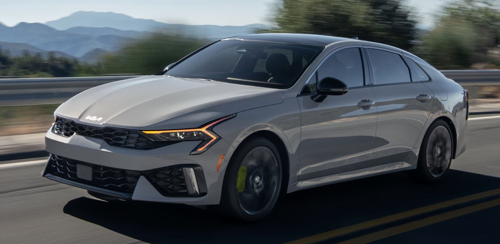 2024 Kia K5 in motion on a scenic road with mountains in the background, showcasing its sleek design.