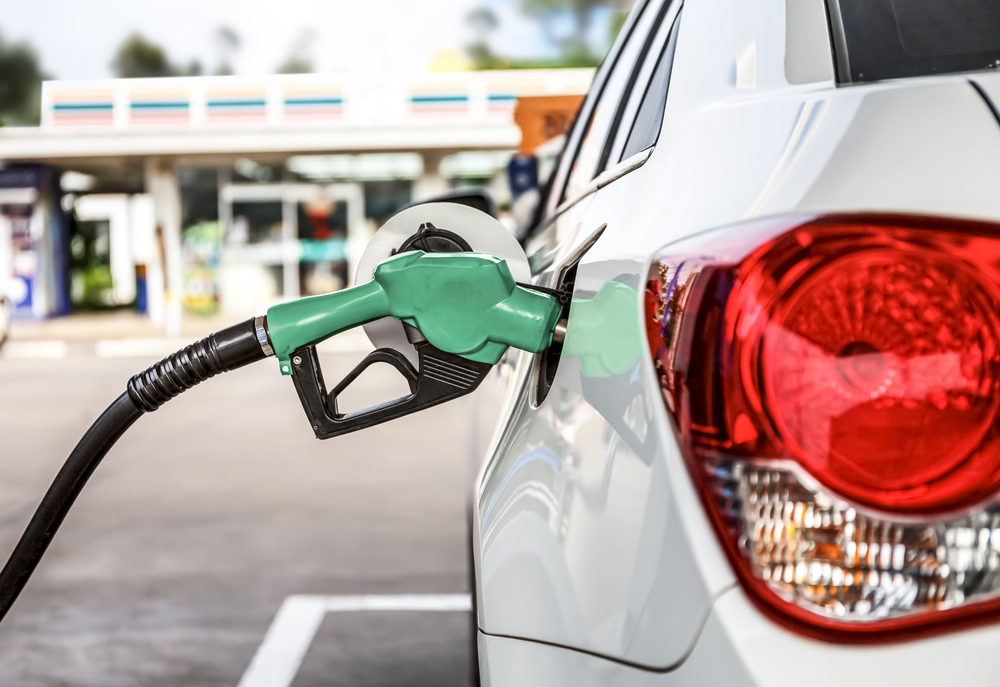 Green gas nozzle is inserted into a white cars fuel tank opening,  a gas station is visible blurred out in the background