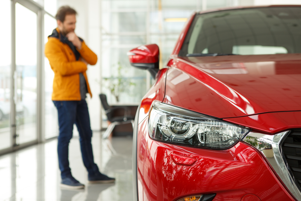 man in background, standing behind a red car thinking as he looks at the car inside of a dealership; man is out of focus in background, with red car being the main focus of photo