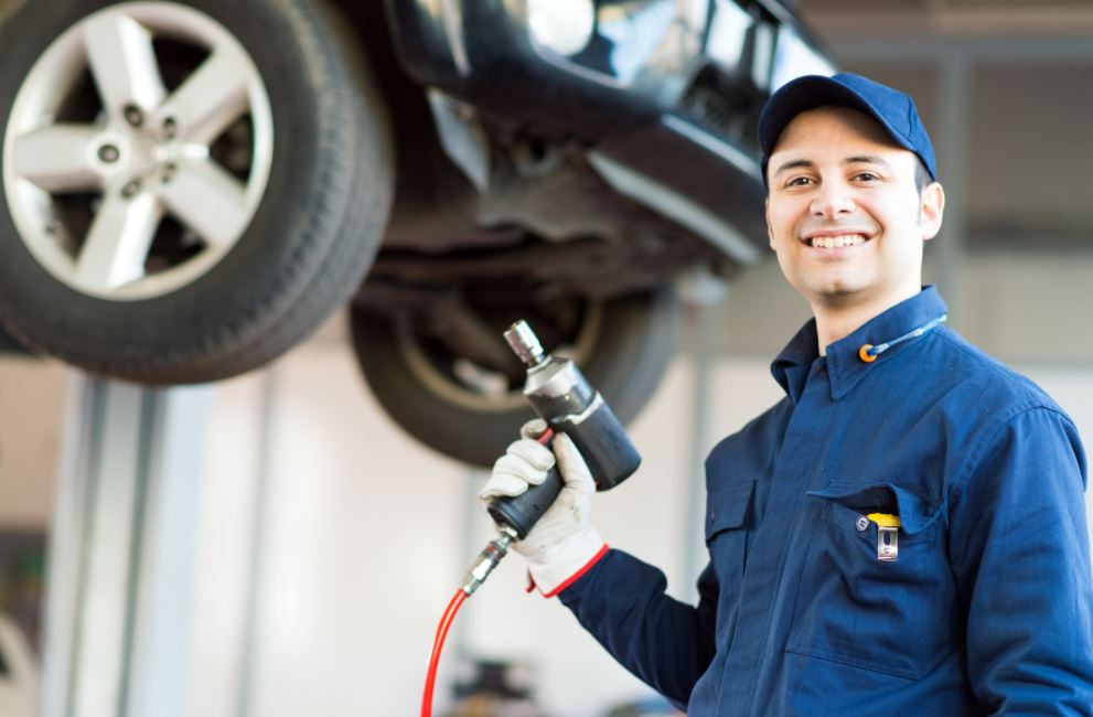 Smiling mechanic holding tool, in the background a car is lifted up on mechanic lift