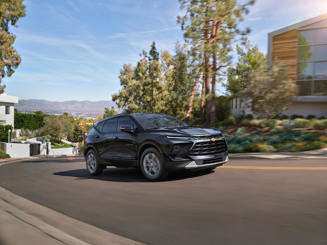 3/4 view of Black 2024 Blazer driving down road in front of houses. 