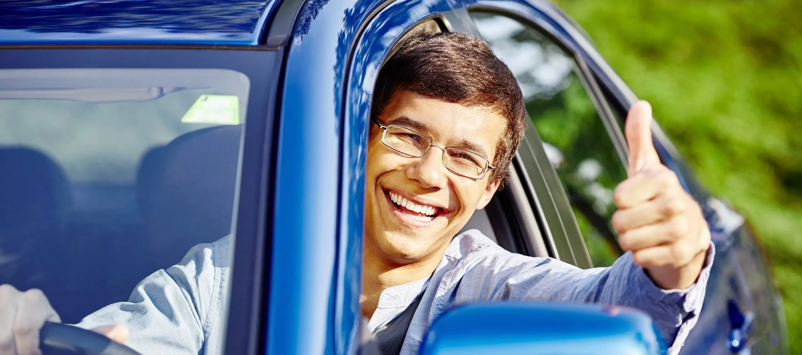 A teenager is sitting in a blue car with his hand and head out the window, he is smiling and giving a thumbs up 