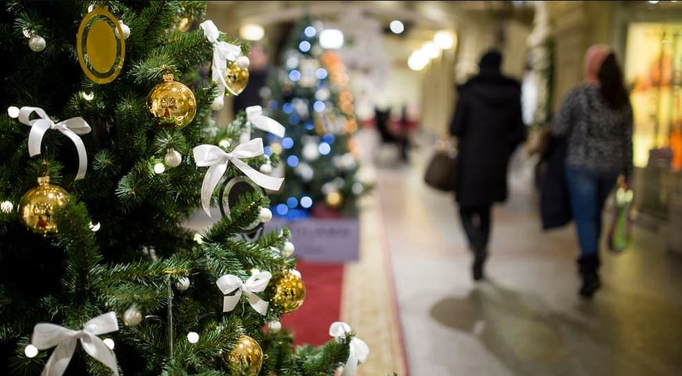 (in focus) a mall's christmas tree decorated with gold ornaments and white ribbon bows, (out of focus) people shopping in the mall