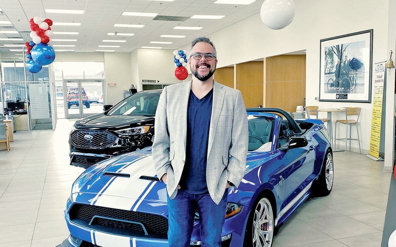 Scott Kunes, Chief of Operations at Kunes Auto Group, pictured inside of Delavan Ford Dealership pased in front of a blue mustang with a white racing stripe on the middle of the hood