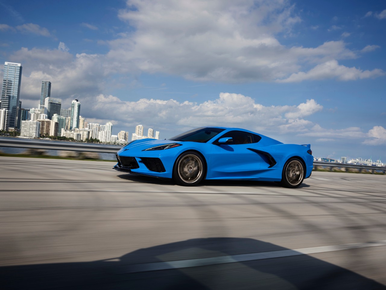 Side view of Blue Chevrolet Corvette Stingray driving in front of city skyline. 
