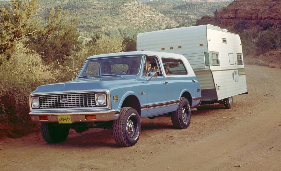 blue vintage 70s Chevy SUV towing a camper on a dirt road