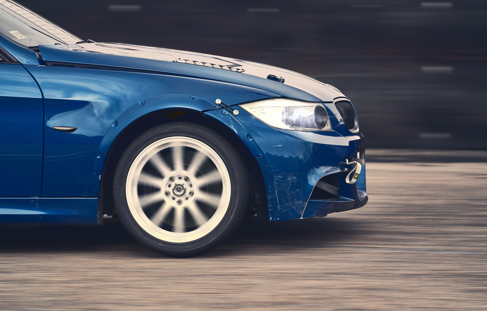 profile view of the passenger side of a blue car as it drives at high speed; focus is on the wheel and tire