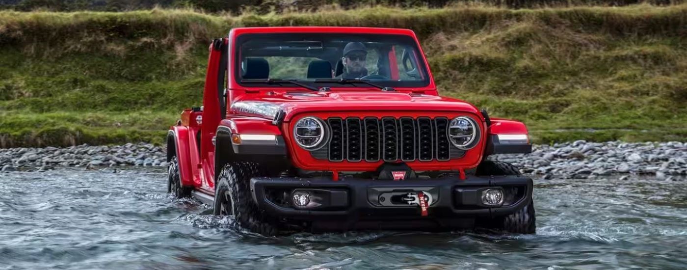 A red 2024 Jeep Wrangler for sale is shown driving through water.