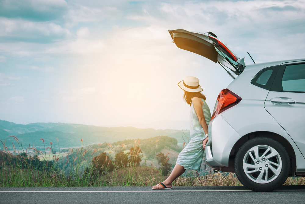 woman with a sunhat sits in the trunk of her white hatchback, parked on the side of the road, as she gazes out into the countryside fields