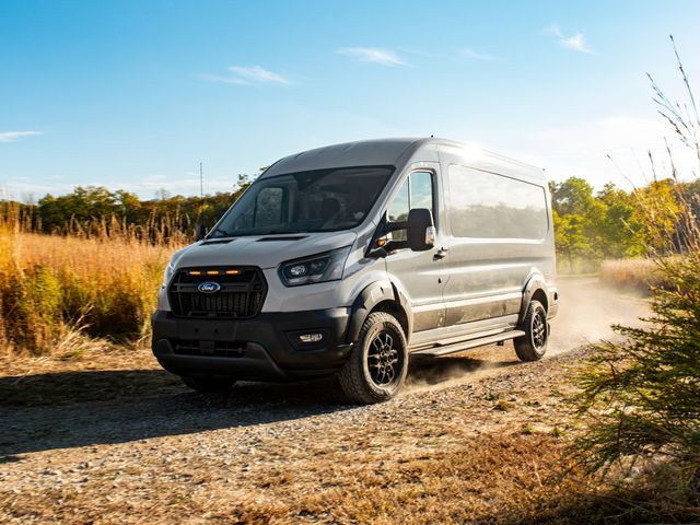 A gray Ford Transit 350 going down a dirt country road with overgrowth on both side