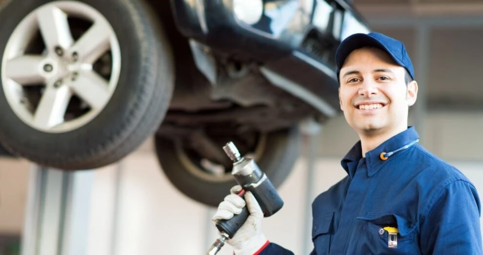 Bottom of Car is up on forklift, Mechanic stands closer to frame smiling wearing a blue jumpsuit holding a tool