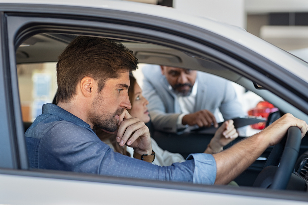 man and spouse thinking inside of a car while a salesman peaks through passenger window with clipboard in hand
