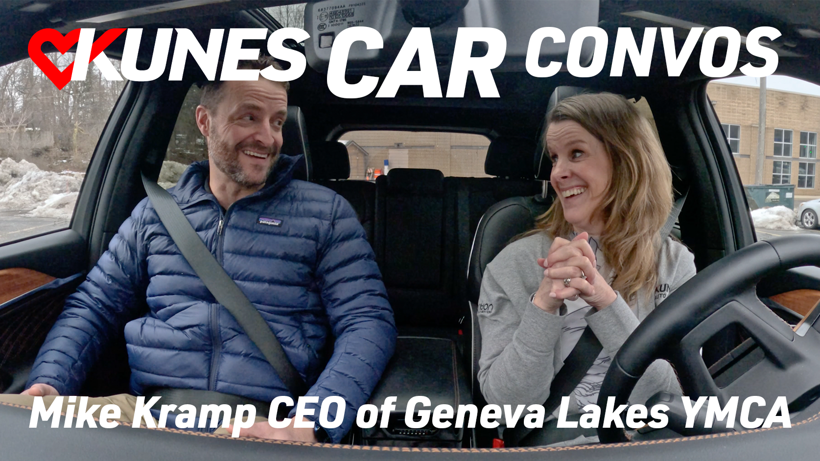 Text reads: Kunes Car Convos; Mike Kramp CEO of Geneva Lakes YMCA
Photo: Mike Kramp, CEO of Geneva Lakes YMCA, and Jen Myers, Marketing Director of Kunes Auto & RV Group, smile at each other while driving in a 2023 Grand Cherokee 4XE