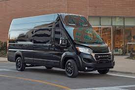 a black ram promaster 3500 Window Van in a passenger side view driving down a main street in a small  town