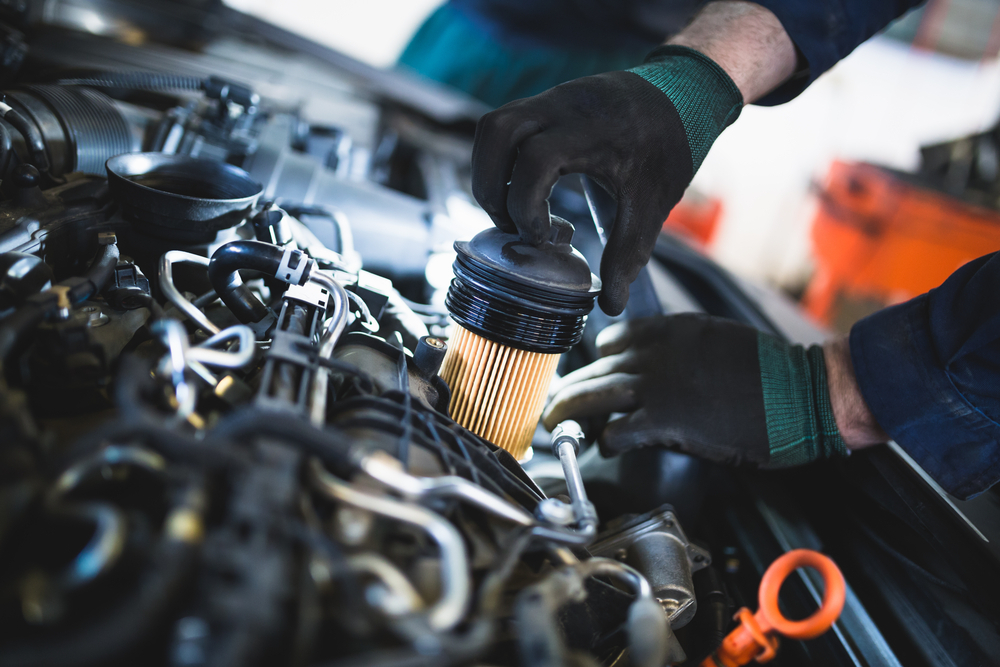 The cars engine is in focus with a pair of hands donning black gloves is installing an air filter into the engine, blurred out in the background is a red tool bench