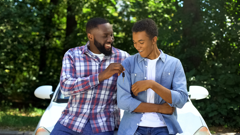 A Father and Son are sitting on the hood of a white car with numerous green trees in the background, the father is handing the son keys to a car and the son is taking them with a smile on his face