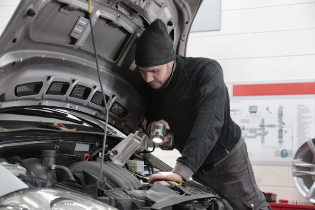 Mechanic in black outfit shining flashlight to inspect under hood of car