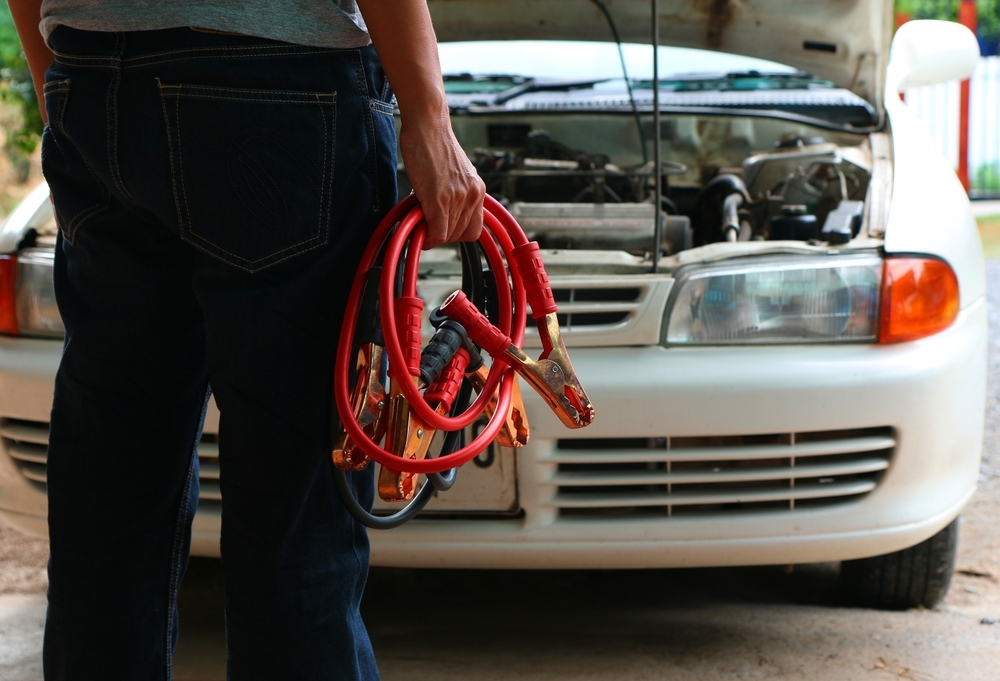 A man is in frame holding jumper cables, only the bottom half of his torso is showing, the jumper cables are red with orange clips, the man is standing in front of a white car with its hood open.