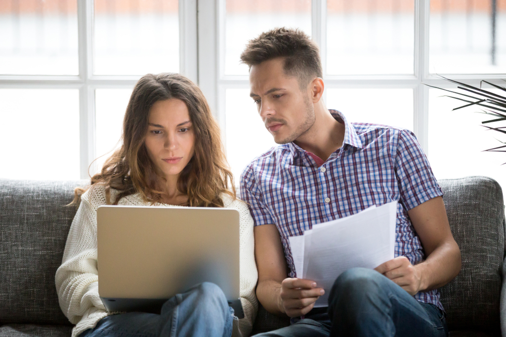 A Couple is sitting on their couch, women on the left man on the right, they are both looking at paperwork, a window is behind them letting in sunlight, on the far left we can see a few leaves of a plant