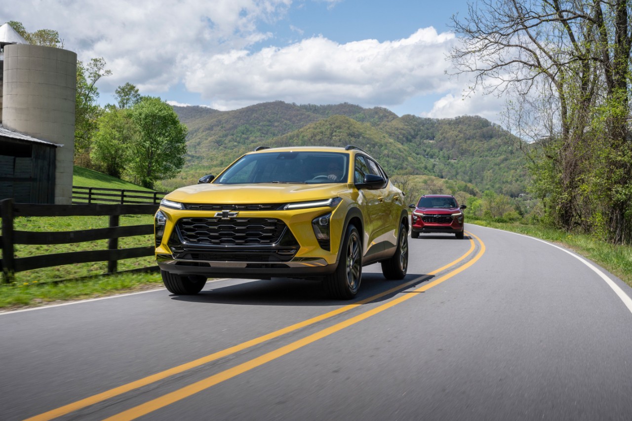 Front 3/4 view of 2024 Chevrolet Trax ACTIV in Nitro Yellow Metallic and RS in Crimson Metallic driving on a road with mountains in the horizon.