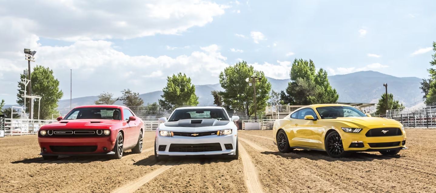 red Dodge Challenger, white wih black hood Chevrolet Camaro, yellow Ford Mustang; parked on a dirt race track with mountains in the background