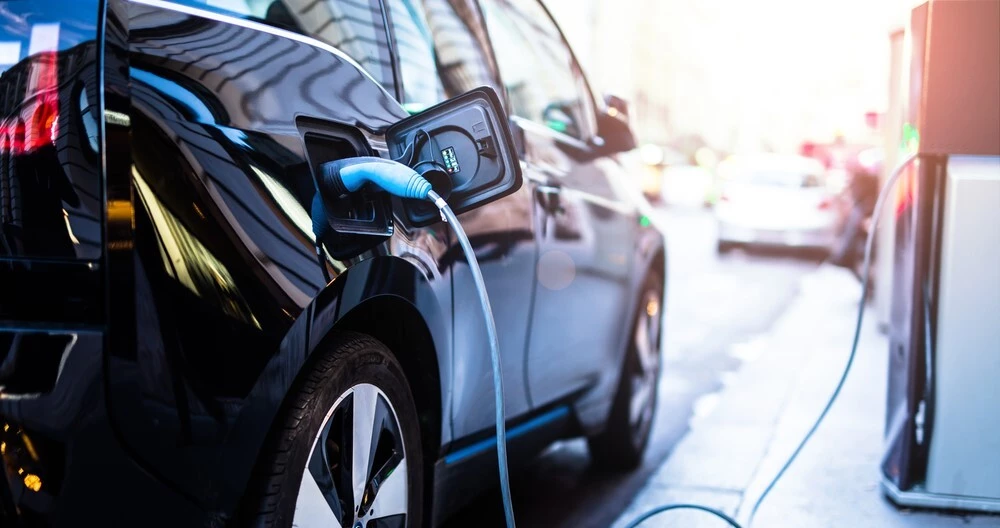 Black Electric Car with a Green EV charger inserted into it, a silver car can be seen blurry in the background