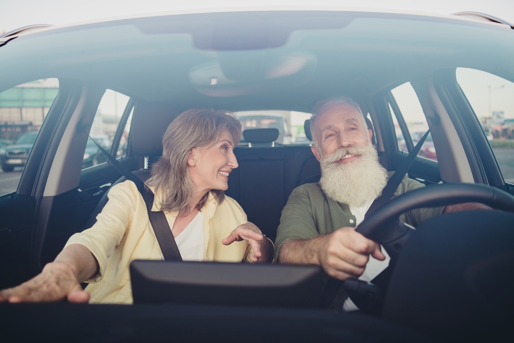 older woman points to infotainment center while smiling at her husband as he drives their vehicle