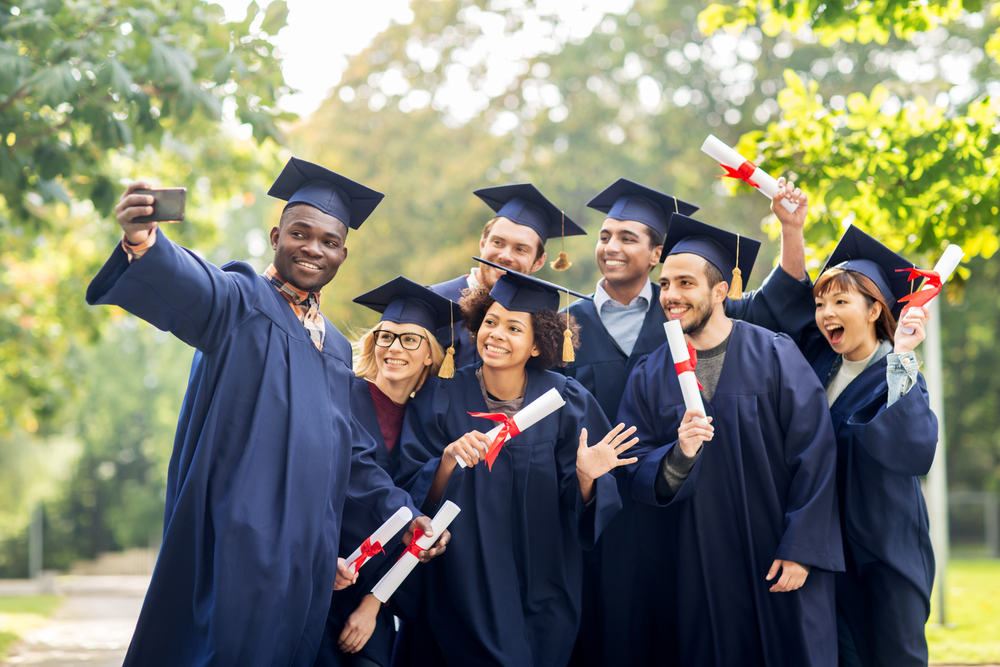 graduates pose for a selfie with their diplomas in their caps and gowns